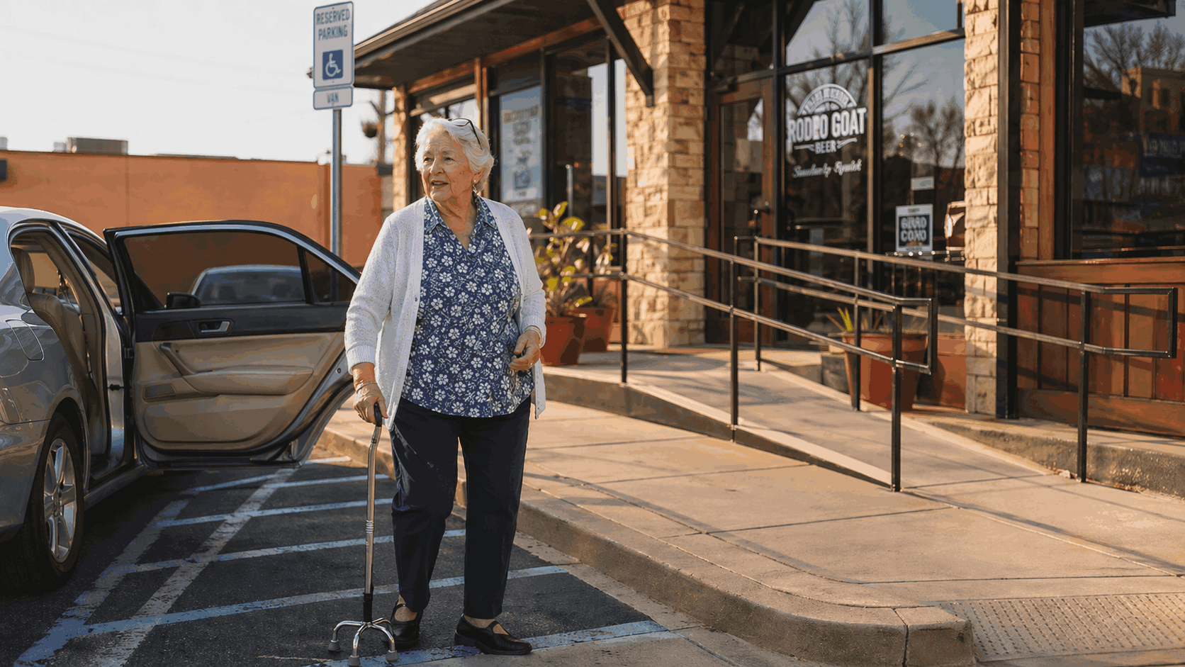 Older Texas woman using a four-legged cane walks from an accessible parking space into a local San Antonio business in late-afternoon light.